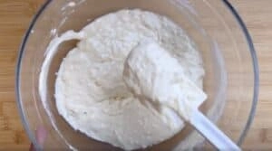 Mixing bread dough in a glass bowl with a spatula, on a wooden surface.
